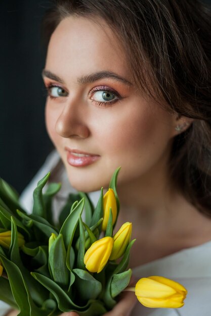 Portrait de printemps de belle jeune femme aux yeux bleus et aux cheveux bouclés avec bouquet de tulipes jaunes sur fond noir Modèle heureux en jeans et chemise blanche Fête des mères