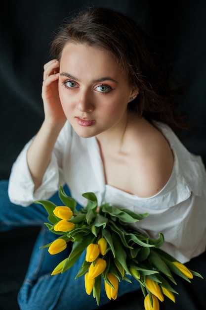 Portrait de printemps de belle jeune femme aux yeux bleus et aux cheveux bouclés avec bouquet de tulipes jaunes sur fond noir Modèle heureux en jeans et chemise blanche Fête des mères