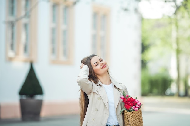 Portrait de printemps d'une belle jeune femme heureuse de 28 ans aux longs cheveux bien coiffés tient un sac en osier dans ses mains avec un bouquet de tulipes dans une rue de la ville Modèle élégant dans un trench-coat