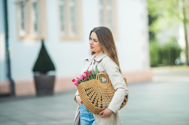 Portrait de printemps d'une belle jeune femme heureuse de 28 ans aux longs cheveux bien coiffés tient un sac en osier dans ses mains avec un bouquet de tulipes dans une rue de la ville Modèle élégant dans un trench-coat