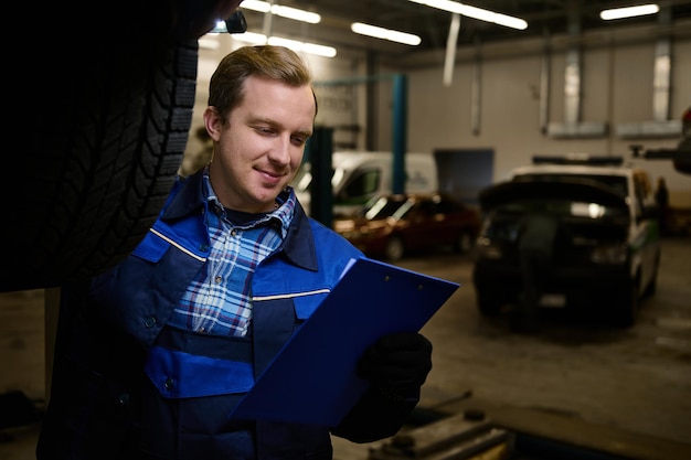 Portrait professionnel d'un bel homme caucasien, mécanicien automobile, technicien faisant une liste de contrôle sur le presse-papiers tout en inspectant la voiture soulevée dans l'ascenseur de l'atelier de réparation. Concept d'entretien de voiture