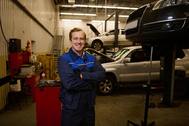 Portrait professionnel confiant d'un beau mécanicien automobile en uniforme souriant avec un sourire à pleines dents, debout dans un garage de service automobile avec une voiture levée en arrière-plan