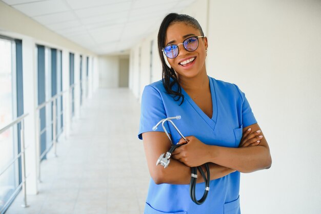 Portrait d'une professionnelle de la santé féminine souriante et confiante avec une blouse de laboratoire, les bras croisés tenant des lunettes. Fond de clinique d'hôpital isolé.
