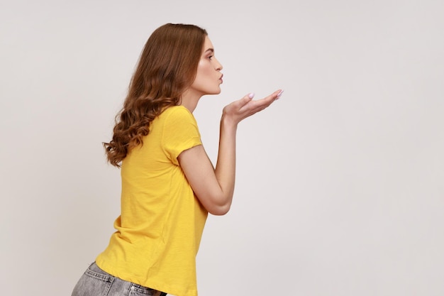 Portrait de profil d'une adolescente aux cheveux ondulés envoyant un baiser aérien avec des lèvres séduisantes, flirtant, exprimant l'affection amoureuse et les sentiments romantiques. Tourné en studio intérieur isolé sur fond gris.