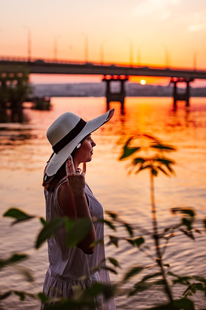 Portrait de profil d'une charmante jeune fille tenant ses mains par le bord d'un chapeau de paille sur une plage fluviale avec...