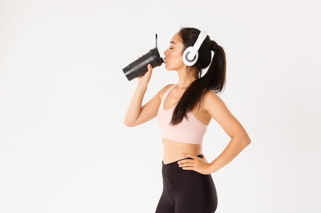 Portrait De Profil D'entraîneur De Fitness Asiatique Sexy, Femme Dans Les écouteurs De L'eau Potable De La Bouteille Pendant L'entraînement Dans La Salle De Gym, Debout