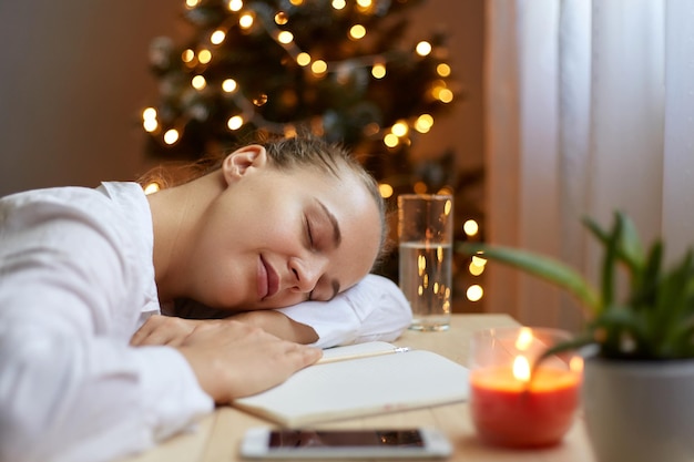 Portrait de profil d'une femme détendue portant une chemise blanche allongée sur une table dans une pièce décorée près de l'arbre de Noël avec son bloc-notes en papier et son téléphone portable se sent somnolent après une sieste