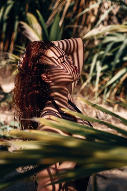 Photo portrait de profil: gros plan d'une belle fille rousse posant dans les branches de palmiers.