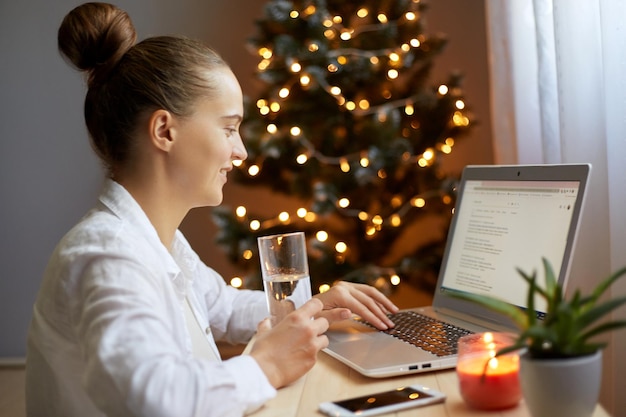 Portrait de profil d'une jolie femme satisfaite avec une coiffure chignon tapant sur un clavier d'ordinateur tout en travaillant à l'intérieur d'un bureau décoré pour les vacances de Noël tenant un verre d'eau et buvant