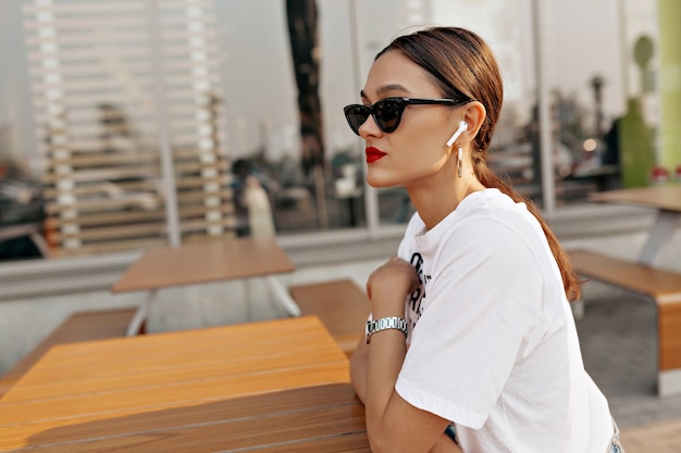 Portrait de profil d'une jolie jolie fille aux cheveux noirs et aux lèvres rouges porte des lunettes et un t-shirt blanc est assis à l'extérieur au soleil