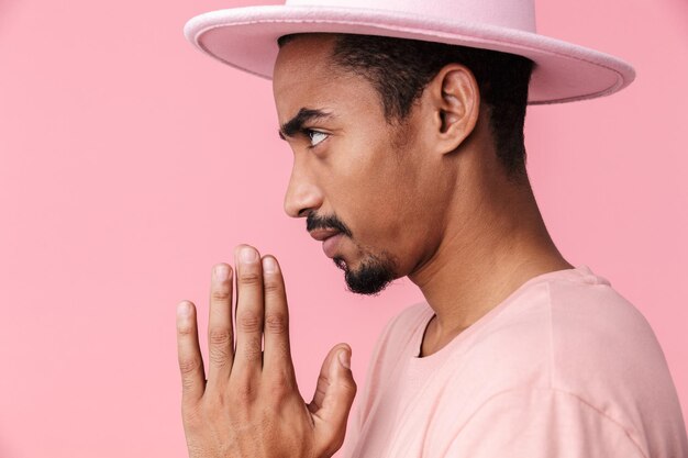 Portrait de profil de la pensée d'un homme afro-américain portant un chapeau regardant de côté et tenant les paumes ensemble isolées sur rose