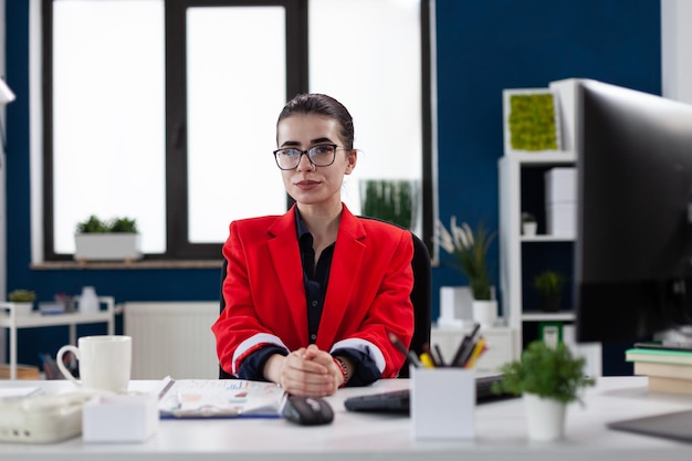 Photo portrait d'un propriétaire de petite entreprise avec des lunettes au bureau de démarrage. employé professionnel en veste rouge qui a l'air confiant dans l'espace de travail de l'entreprise. femme d'affaires confiante posant assis au bureau.