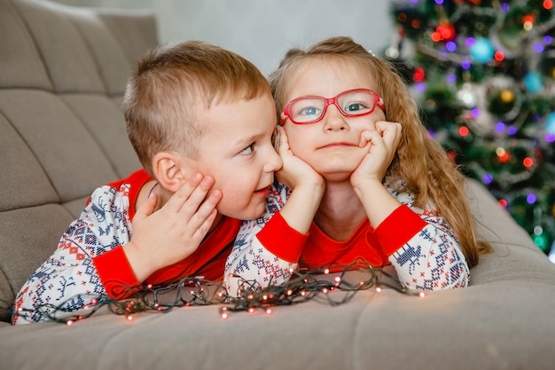 Portrait de quatre ans - frère et soeur jumeaux en pyjama à la maison sur le canapé avec arbre de Noël