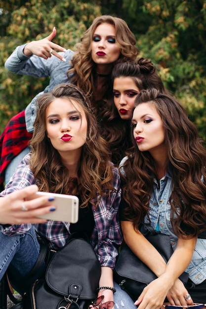 Portrait de quatre copines portant des vêtements décontractés à la mode, des coiffures et du maquillage posant tout en faisant un autoportrait via un téléphone portable assis à l'extérieur dans le parc le jour.