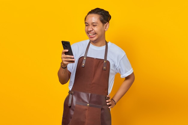 Portrait de rire beau jeune homme asiatique portant un tablier regardant un message sur un smartphone