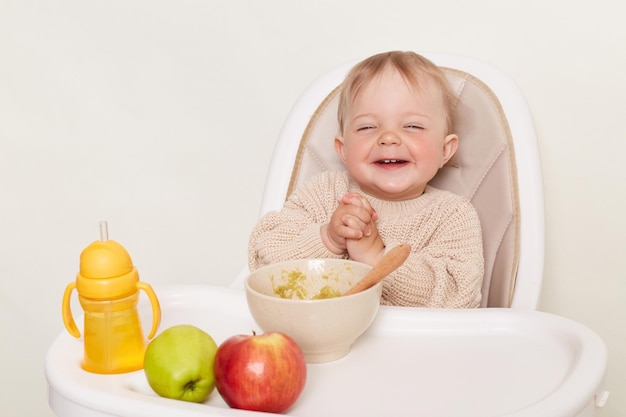 Portrait de rire bébé drôle portant un pull beige assis dans une chaise d'enfant s'amusant pendant le dîner garde les mains ensemble isolé sur fond blanc