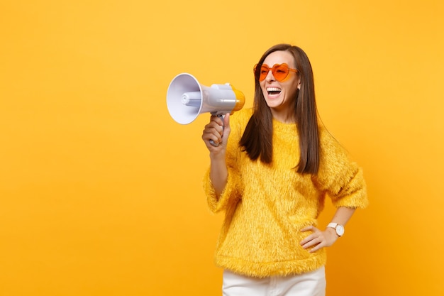 Portrait de rire joyeuse jeune femme en pull de fourrure, lunettes coeur orange tenant un mégaphone isolé sur fond jaune vif. Les gens émotions sincères, concept de style de vie. Espace publicitaire.