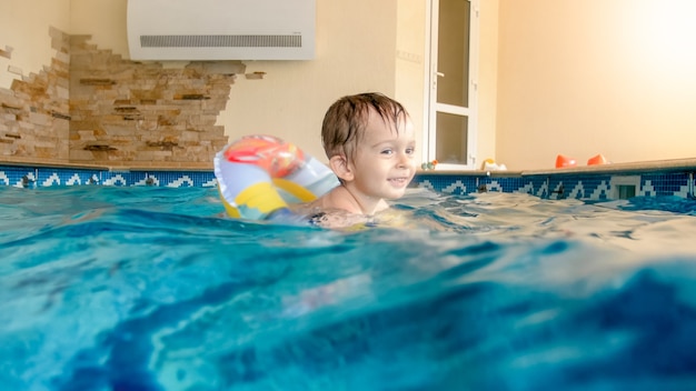 Portrait de rire et de sourire petit garçon de 3 ans nageant avec un anneau gonflable coloré et jouant avec un ballon de plage dans la piscine