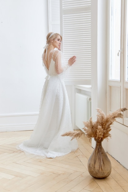 Portrait romantique d'une femme près de la fenêtre dans une belle longue robe blanche à la maison. La fille est blonde aux yeux bleus et beau maquillage sur son visage. Cosmétiques naturels