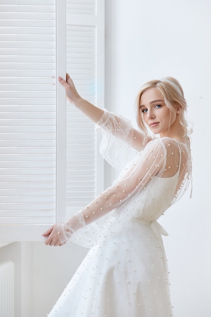 Portrait romantique d'une femme près de la fenêtre dans une belle longue robe blanche à la maison.