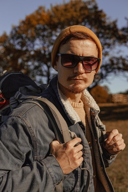 Portrait d'un routard masculin portant des lunettes de soleil avec un sac de voyage regardant la caméra dans la forêt d'automne