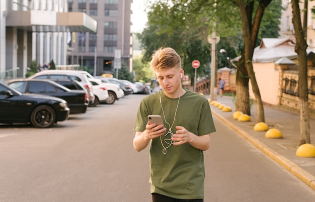 Portrait de rue d'un beau jeune homme écoute de la musique dans des écouteurs et utilise un smartphone