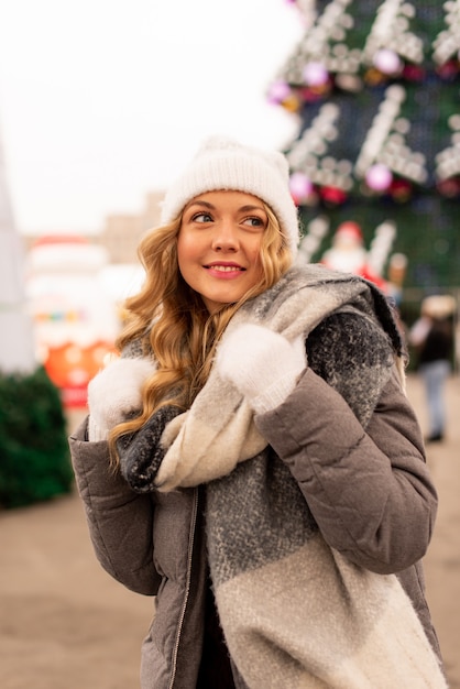 Portrait De Rue De La Belle Jeune Femme Souriante Sur La Foire De Noël. Dame Portant Des Vêtements Tricotés D'hiver Classiques Et élégants. Modèle Regardant La Caméra. Effet De Neige Magique. Fermer