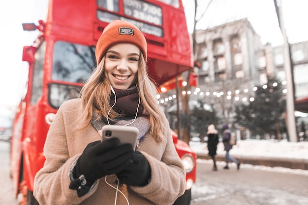 Portrait de rue d'une douce dame vêtue d'un chapeau et d'un manteau, écoute de la musique dans les écouteurs, tient un smartphone dans ses mains et regarde la caméra