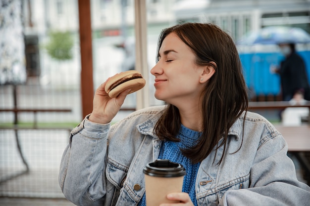 Portrait de rue d'une jeune femme joyeuse appréciant un hamburger et un café à l'extérieur.