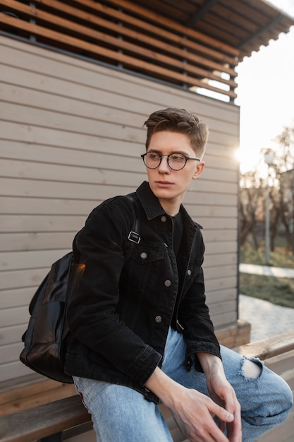 Portrait de rue jeune homme américain avec des lunettes vintage en veste en jean avec sac à dos en ville au coucher du soleil