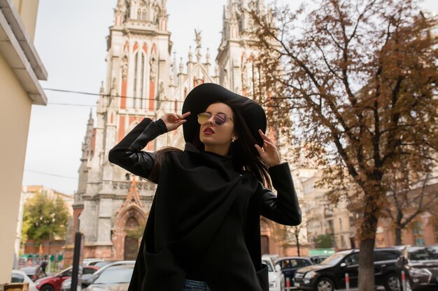 Photo portrait de rue d'un modèle attrayant en manteau noir tendance et chapeau élégant. concept de mode féminine