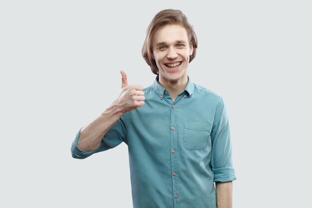 Portrait de satisfait heureux beau jeune homme blond aux cheveux longs en chemise décontractée bleue debout avec les pouces vers le haut et sourire à pleines dents à la caméra. tourné en studio intérieur, isolé sur fond gris clair.