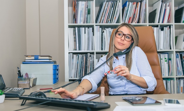 Portrait d'une secrétaire blonde non motivée polissant les ongles sur le lieu de travail tout en parlant au téléphone