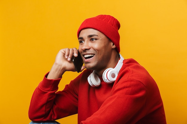 Portrait d'un séduisant jeune homme africain décontracté et heureux assis sur un sol sur un mur jaune, parlant au téléphone portable