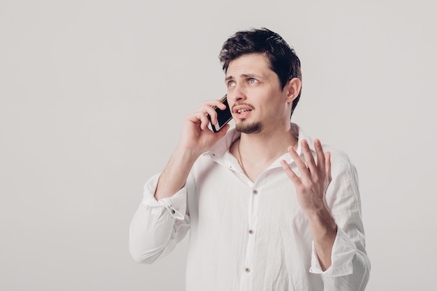 Portrait de séduisante jeune homme brune en chemise blanche parlant sur le smartphone sur fond gris. Lumière douce