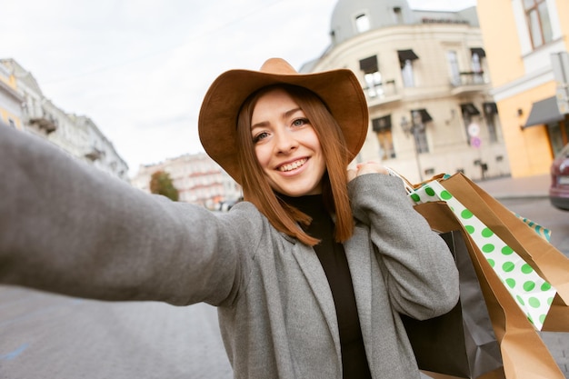 Portrait de selfie. Jolie jolie femme accro du shopping heureuse en manteau d'automne et chapeau tenant de nombreux sacs en papier dans la ville européenne
