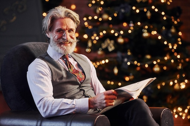 Le portrait d'un senior élégant aux cheveux gris et à la barbe est dans une salle de noël décorée avec un livre dans les mains.