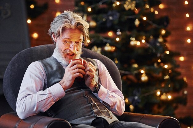 Le portrait d'un senior élégant aux cheveux gris et à la barbe est dans une salle de noël décorée avec une tasse de boisson.