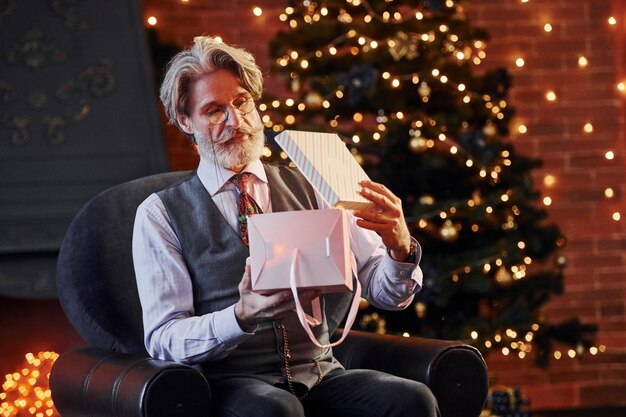 Le portrait d'un senior élégant aux cheveux gris et à la barbe se trouve dans une salle de noël décorée avec une boîte-cadeau.