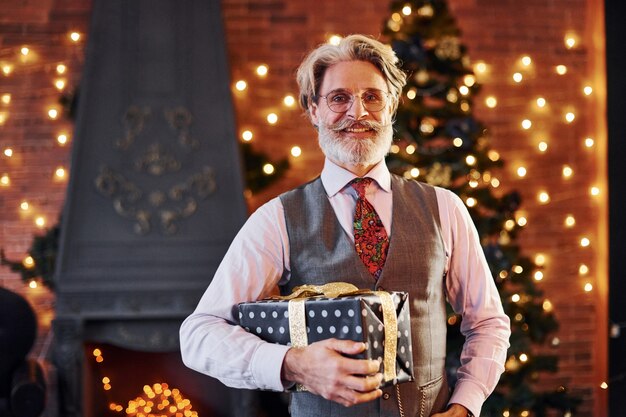 Portrait d'un senior élégant aux cheveux gris et à la barbe en veste et chemise blanche se trouve dans une salle de noël décorée.