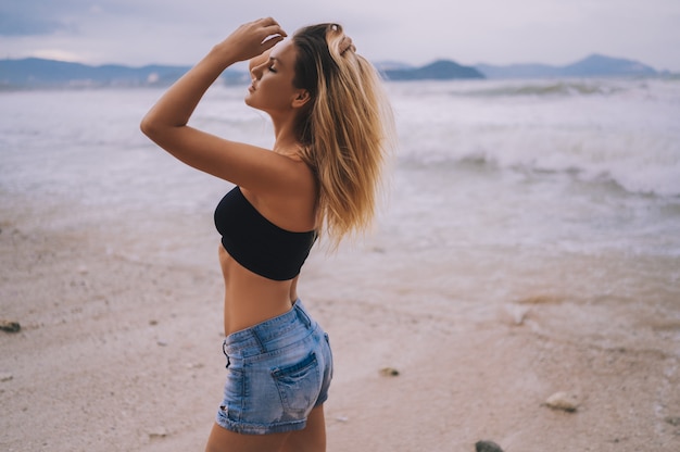 Portrait sensuel de blonde jeune femme européenne debout et posant sur la plage à l'océan de la mer orageuse. Vagues fortes et vent dans les cheveux. Voyager le long de l'Asie, concept de mode de vie actif