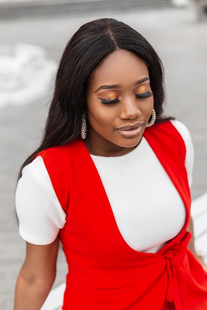 Portrait sensuel sexy jeune femme noire avec de belles lèvres avec une peau propre et saine avec des cheveux longs en gilet rouge tendance en T-shirt blanc dans la rue. Jolie fille assise avec les yeux fermés à l'extérieur.