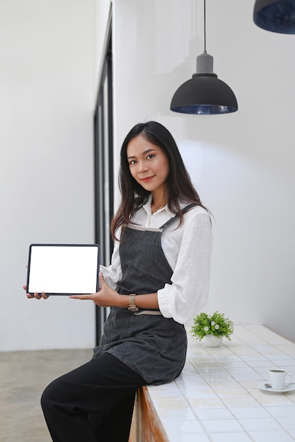 Portrait d'une serveuse sympathique tenant et montrant une tablette numérique avec un écran vide.