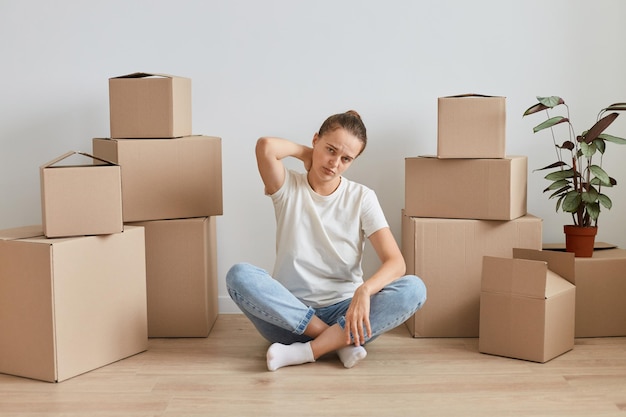 Portrait si malade fatigué femme portant un T-shirt blanc assis sur le sol près de boîtes en carton avec pile personnelle, étant fatigué lors de l'emménagement dans une nouvelle maison, ressentant des douleurs dans le cou.