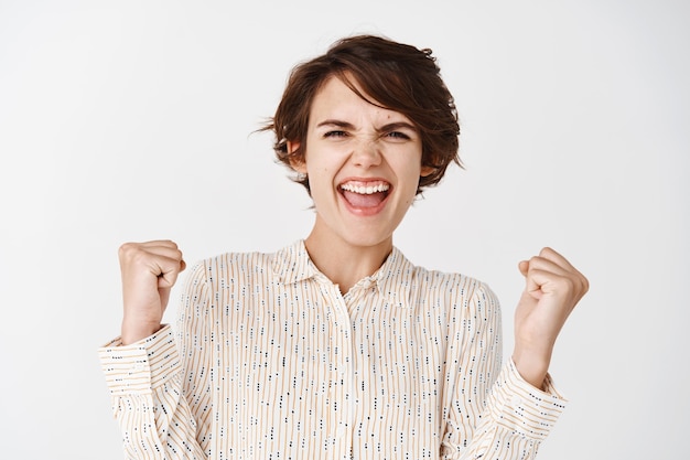 Portrait sincère d'une femme heureuse remportant un prix, se réjouissant et criant oui de joie, faisant une pompe à poing pour célébrer la victoire, triomphant sur un mur blanc