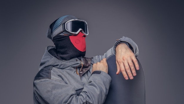 Portrait d'un snowboardeur vêtu d'un équipement de protection complet pour le snowboard extrême posant avec le snowboard dans un studio. Isolé sur un fond gris.