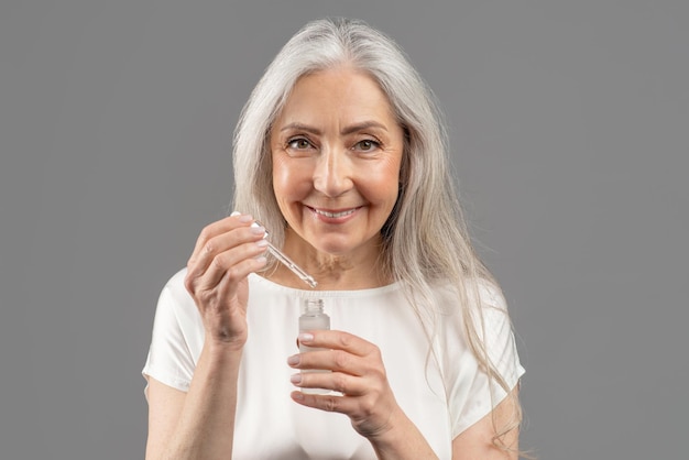 Portrait de soins de la peau et de bien-être d'une femme âgée souriante appliquant du sérum pour le visage avec un compte-gouttes sur gris