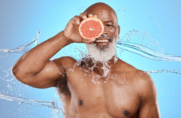 Portrait de soins de la peau et homme senior avec pamplemousse en studio pour les éclaboussures d'eau de beauté et le bien-être sur fond bleu Éclaboussure de beauté du visage sur le produit de fruits par un homme âgé pour la peau du visage et le nettoyage