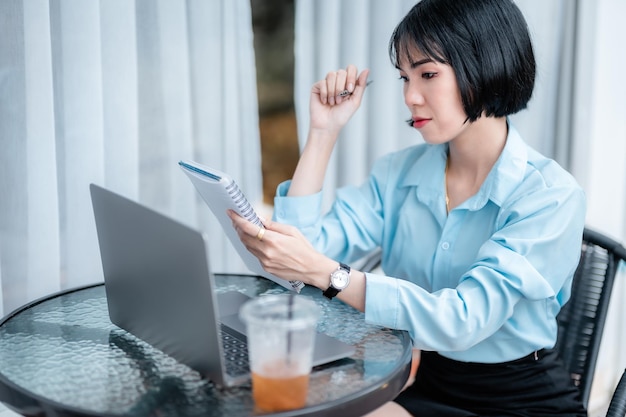 Un portrait de souriant avec des appareils dentaires de femmes d'affaires indépendantes asiatiques écrit des notes sur un ordinateur portable travaillant avec un ordinateur portable avec une tasse de café et un smartphone dans un café