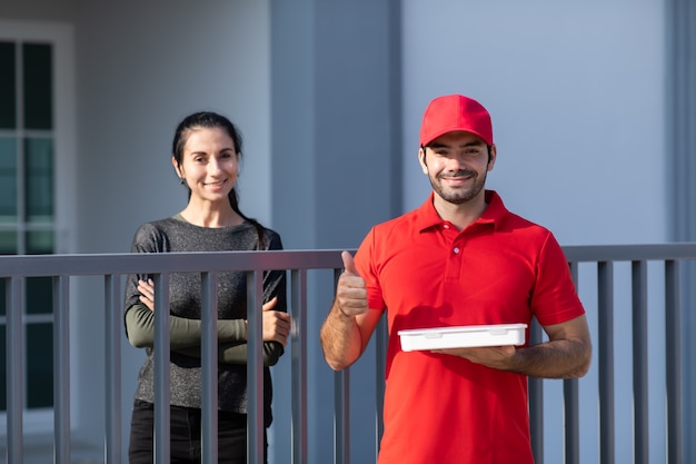 Portrait Souriant jeune livreur en uniforme rouge tenant une boîte devant la maison.